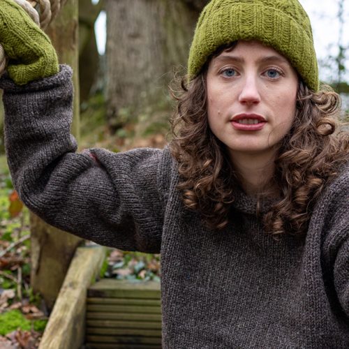 Woman wearing a wool natural sweater with green wollen hand warmers and hat under a tree