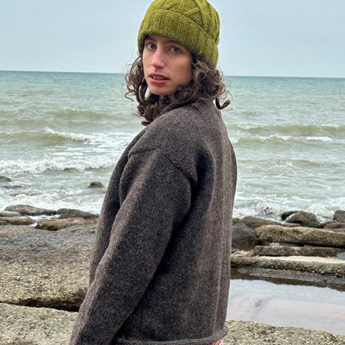 Woman wearing a pure wool natural jumper, with moss green hand warmers and hat on a winter beach